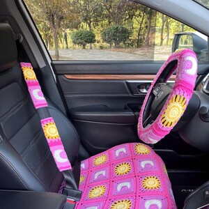 May include: A pink, yellow, and white crochet car seat cover, steering wheel cover, and seat belt covers. The seat cover features a square pattern with yellow sunflowers and white crescent moon shapes. The steering wheel cover and seat belt covers have a similar design.