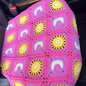 May include: Handmade crochet car seat cover featuring a vibrant floral design. The cover is primarily pink, with yellow and white accents forming sun and crescent moon patterns. The squares are approximately 6 inches by 6 inches.