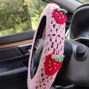 May include: Pink crocheted steering wheel cover with strawberry appliques. The cover is made of a soft, textured yarn and features a delicate crochet pattern.