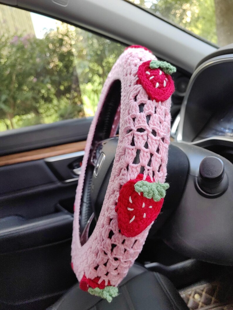 May include: Pink crocheted steering wheel cover with strawberry appliques. The cover is made of a soft, textured yarn and features a delicate crochet pattern.