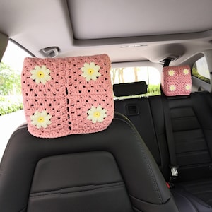 May include: Two pink crocheted car headrest covers with yellow daisy flowers. The headrest covers are in the back seat of a car.