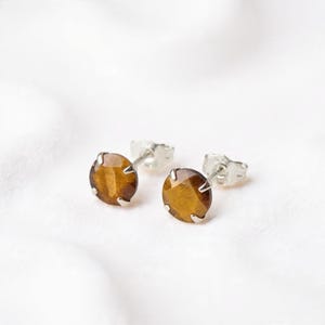 May include: A pair of round stud earrings featuring faceted tiger eye gemstones set in silver-toned metal prongs. Each earring has a metallic post back closure, showcasing the natural brown and golden chatoyant bands of the stone.