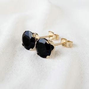 May include: A pair of stud earrings featuring round, faceted black stones set in gold-toned metal prongs. Each earring includes a thin gold-toned post and a matching butterfly-style earring back, showcasing a classic design with a polished finish.