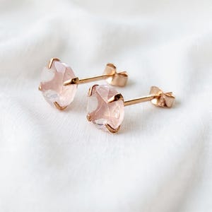 Può includere: Un paio di orecchini a lobo con pietre preziose rotonde e sfaccettate color rosa pallido, incastonate in griffe di metallo dorato. Ogni orecchino presenta un sottile perno dorato e una chiusura a farfalla abbinata, con superficie lucida.