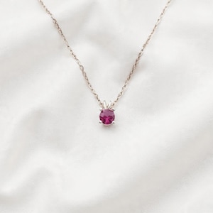 May include: A silver necklace with a small, round, magenta-colored gemstone pendant. The pendant is set in a four-prong setting and hangs from a delicate chain. The necklace is displayed on a white, textured background.