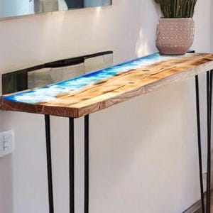 Puede incluir: Una mesa consola de madera con una tapa de resina epoxi azul y patas de horquilla negras. La mesa está contra una pared blanca y tiene una pequeña planta en una maceta encima.