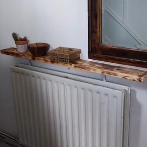 May include: A white radiator with a wooden shelf above it. The shelf is decorated with a small potted plant, a wooden bowl, and a woven basket. The wall is painted white.