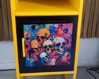 May include: A yellow metal nightstand with a black door featuring a colorful skull graphic. The nightstand has a shelf and a clock on top.