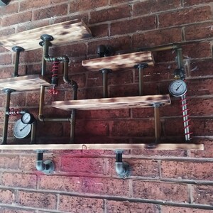 Può includere: Un'unità a scaffale in legno rustico con un design steampunk. Gli scaffali sono realizzati in legno riciclato e sono sostenuti da tubi e raccordi industriali. L'unità presenta due manometri e una molla rossa.