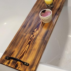 May include: A wooden bath tray with black metal handles. The tray has a burnt wood finish and is sitting on the edge of a white bathtub. There are two small candles in ceramic holders on the tray.