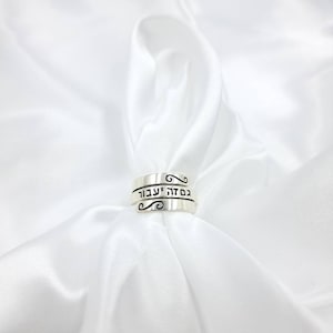 May include: A silver ring with Hebrew writing that reads "Blessed are you, Lord our God, King of the Universe." The ring is on a white fabric background.