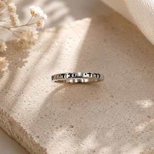 May include: A thin, polished silver-toned metal ring featuring a band engraved with black Hebrew lettering. The ring has a smooth, rounded exterior surface and a consistent width throughout the entire circumference of the circular band.