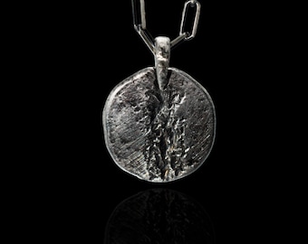 May include: A silver pendant necklace with a round, textured charm. The charm has a rough, organic texture and is suspended from a silver chain.