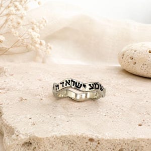 May include: A silver-toned ring with a wavy, irregular band design. The outer surface of the ring features black engraved Hebrew text that reads Shema Yisrael. The metal has a polished finish, and the band has a distinct, non-uniform shape.