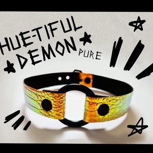 Könnte beinhalten: Ein gold-schwarzer Choker mit einem großen schwarzen Ring. Der Choker hat ein Krokodil-Prägemuster und liegt auf einem weißen Hintergrund. Der Text "HUE-TIFUL DEMON PURE" ist in schwarzem Marker auf dem Hintergrund geschrieben.