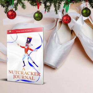 May include: A journal titled "A Nutcracker Journal" with a colorful illustration of a nutcracker soldier. White ballet pointe shoes with ribbons are next to the journal. Christmas ornaments and greenery are in the background.