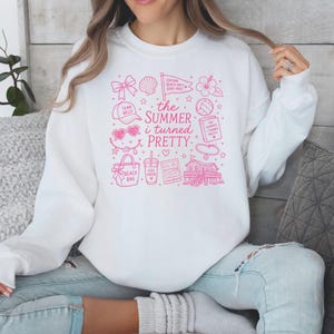 Könnte beinhalten: Ein weißes Sweatshirt mit Rundhalsausschnitt und einem rosa Grafikdruck mit dem Text 'the Summer i turned PRETTY', umgeben von verschiedenen Illustrationen zum Thema Strand, darunter eine Muschel, ein Volleyball, eine Strandtasche und ein Haus.
