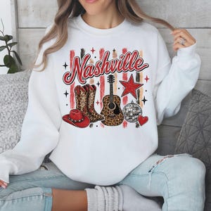 Puede incluir: Una sudadera blanca de cuello redondo con un estampado gráfico que incluye la palabra Nashville en letra roja. El diseño presenta una bota de vaquero con estampado de leopardo, una guitarra acústica de leopardo, una estrella roja, un sombrero rojo y una bola de discoteca.
