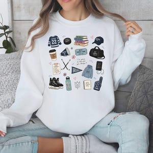 Puede incluir: Sudadera blanca con varias ilustraciones temáticas de hockey, incluyendo una camiseta con el número 22, un disco de hockey, libros, una gorra, una bolsa de deporte, una taza de café, palos de hockey, patines de hielo y un dedo de espuma.