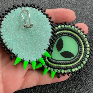 May include: Round, handmade earrings featuring a green alien design. The earrings have a black beaded border, a green center, and neon green spikes. The backs are a glittery, light green fabric. The earrings are held in a hand.