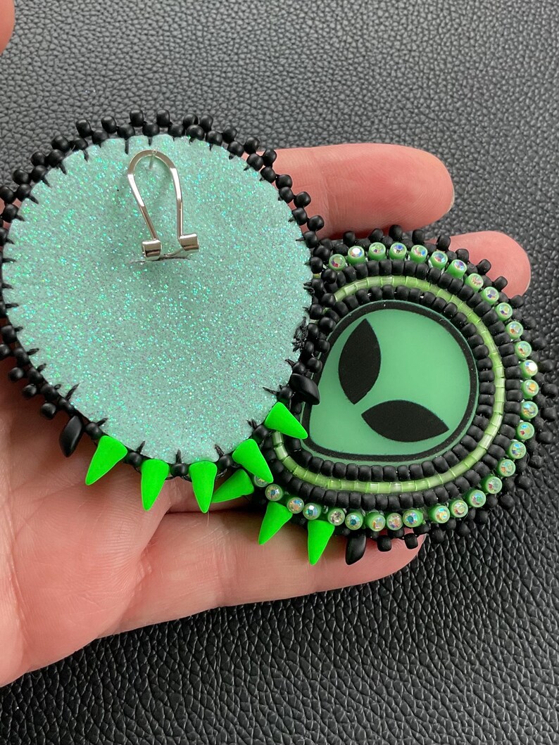 May include: Round, handmade earrings featuring a green alien design. The earrings have a black beaded border, a green center, and neon green spikes. The backs are a glittery, light green fabric. The earrings are held in a hand.