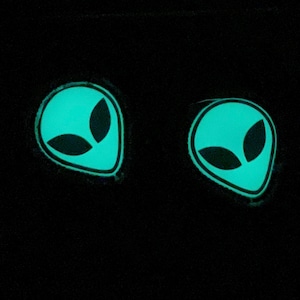 K&ouml;nnte beinhalten: Zwei leuchtend t&uuml;rkisgr&uuml;ne Alien-Kopf-Patches mit schwarzen ovalen Augen und schwarzem Umriss, vor schwarzem Hintergrund. Die Patches haben einen leicht verwitterten Look.