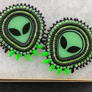 May include: Two handmade alien-themed earrings. Each earring features a light green alien face with black eyes, surrounded by black and green beads. The earrings have bright green spikes and are set in a small box.