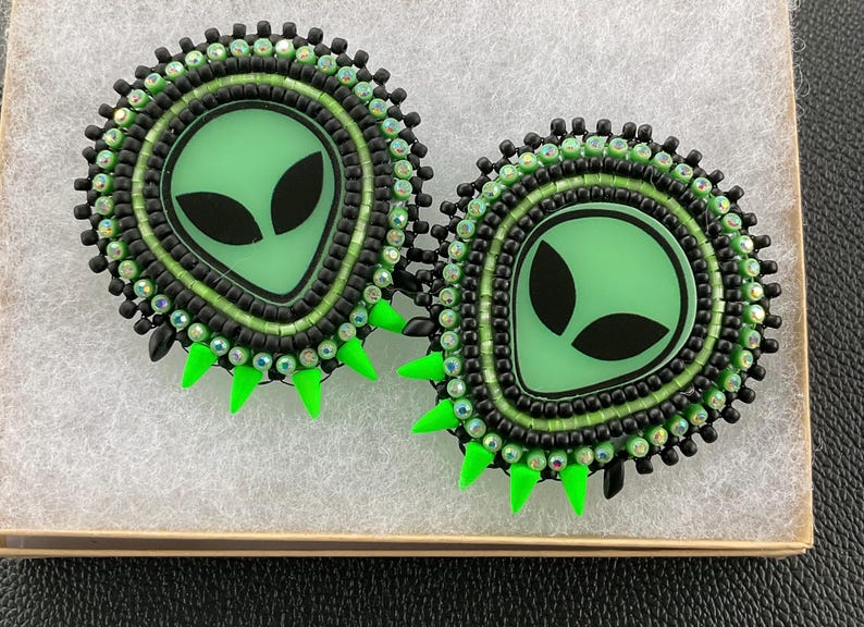 May include: Two handmade alien-themed earrings. Each earring features a light green alien face with black eyes, surrounded by black and green beads. The earrings have bright green spikes and are set in a small box.