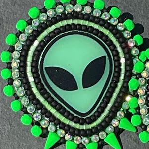 May include: A round, beaded alien face design in shades of green, black, and clear. The alien face is light green with black eyes. The design is surrounded by black beads, clear crystals, and bright green accents.