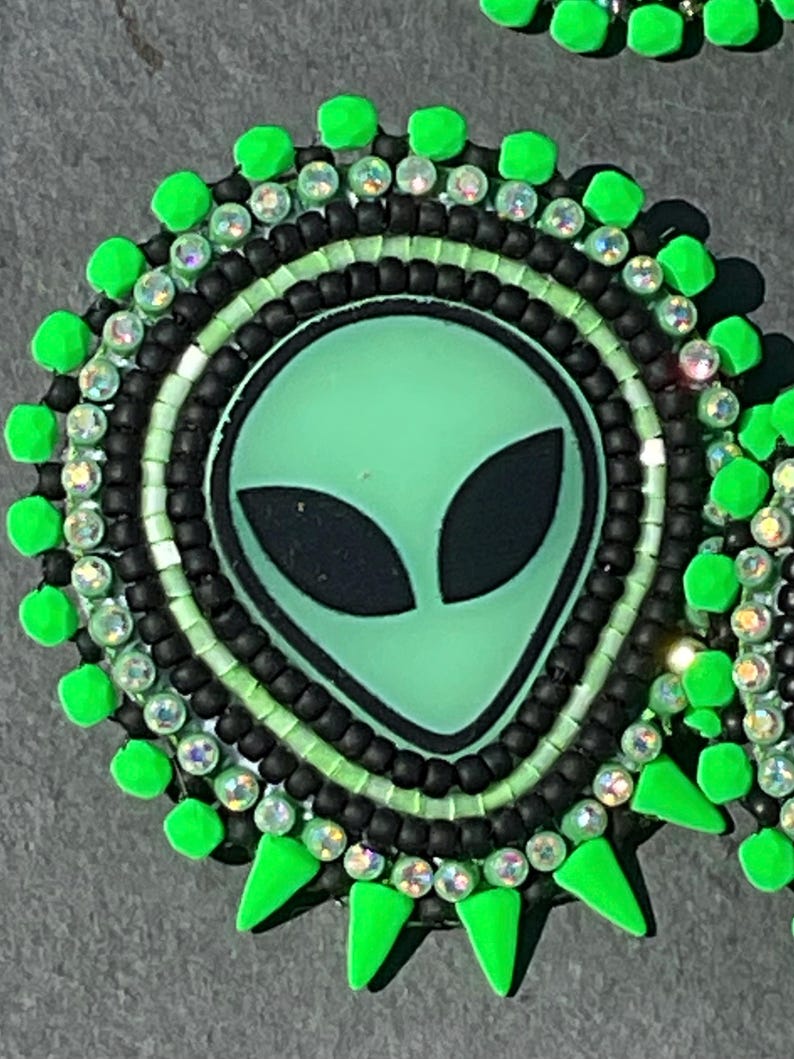 May include: A round, beaded alien face design in shades of green, black, and clear. The alien face is light green with black eyes. The design is surrounded by black beads, clear crystals, and bright green accents.