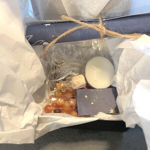 May include: A gift box filled with various items, including a rolled-up blue paper tied with twine, a small glass bottle with a cork, orange beads, a white candle, and a small blue card with gold stars. The box is lined with white tissue paper.