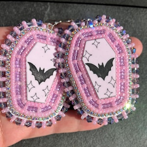 May include: A pair of beaded earrings featuring a central coffin-shaped charm with a light pink background, a black bat silhouette, and star accents. The charm is surrounded by layers of light pink, lavender, and iridescent crystal beads.