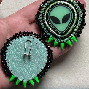 K&ouml;nnte beinhalten: Handgefertigte Ohrringe mit Alien-Motiv. Jeder Ohrring zeigt ein gr&uuml;nes Alien-Gesicht mit schwarzen Augen, umgeben von schwarzen Perlen und gr&uuml;nen Akzenten. Die R&uuml;ckseiten sind hellblau mit einem silbernen Glitzereffekt und einem silbernen Ohrhaken.