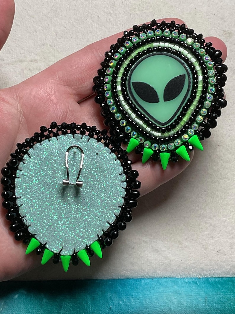 K&ouml;nnte beinhalten: Handgefertigte Ohrringe mit Alien-Motiv. Jeder Ohrring zeigt ein gr&uuml;nes Alien-Gesicht mit schwarzen Augen, umgeben von schwarzen Perlen und gr&uuml;nen Akzenten. Die R&uuml;ckseiten sind hellblau mit einem silbernen Glitzereffekt und einem silbernen Ohrhaken.