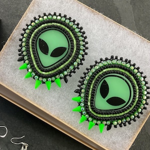 May include: Pair of handmade alien-themed earrings. Each earring features a light green alien face with black eyes, surrounded by black and green beads. Bright green spikes are at the bottom of each earring. The earrings are in a small box.