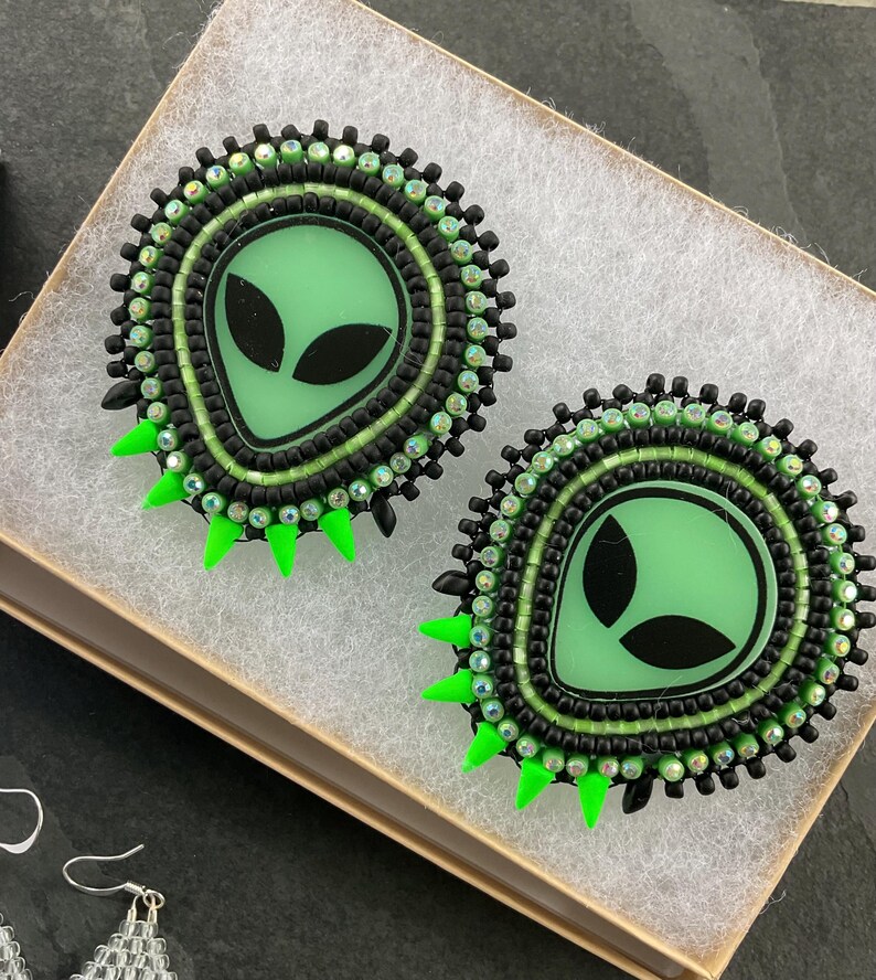 May include: Pair of handmade alien-themed earrings. Each earring features a light green alien face with black eyes, surrounded by black and green beads. Bright green spikes are at the bottom of each earring. The earrings are in a small box.