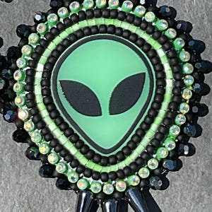 May include: Handmade beaded earrings featuring a green alien face with black eyes. The earrings have a circular design with black, green, and iridescent beads. The bottom of the earrings have black spike-shaped accents.
