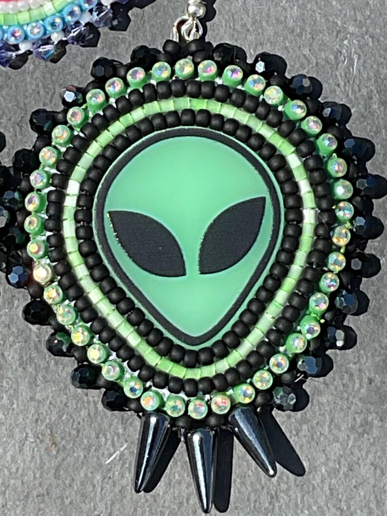 May include: Handmade beaded earrings featuring a green alien face with black eyes. The earrings have a circular design with black, green, and iridescent beads. The bottom of the earrings have black spike-shaped accents.