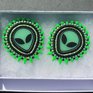 May include: A pair of handmade alien-themed earrings. Each earring features a green alien face with black eyes, surrounded by black and iridescent beads. The outer edge is adorned with bright green spikes and beads, presented in a white box.