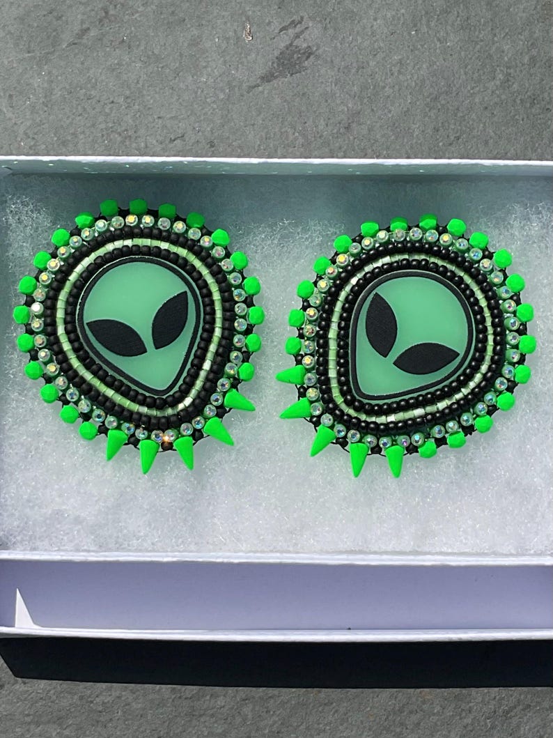 May include: A pair of handmade alien-themed earrings. Each earring features a green alien face with black eyes, surrounded by black and iridescent beads. The outer edge is adorned with bright green spikes and beads, presented in a white box.