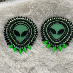 May include: Two beaded alien-themed pins. Each pin features a green alien face with black eyes, surrounded by black and green beads. The pins have a green, spiky bottom edge, and are set against a white, fluffy background.