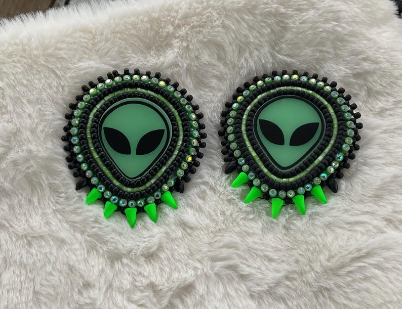 May include: Two beaded alien-themed pins. Each pin features a green alien face with black eyes, surrounded by black and green beads. The pins have a green, spiky bottom edge, and are set against a white, fluffy background.