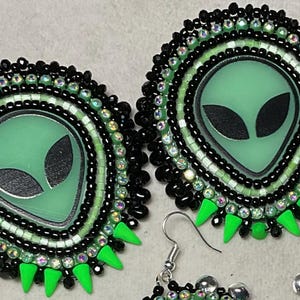 K&ouml;nnte beinhalten: Handgefertigte Ohrringe zum Thema Alien. Jeder Ohrring zeigt ein gr&uuml;nes Alien-Gesicht mit schwarzen Augen, umgeben von schwarzen und irisierenden Perlen. Die Ohrringe haben einen silbernen Haken und gr&uuml;ne dreieckige Akzente.