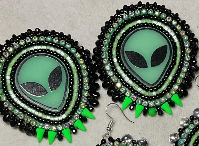 K&ouml;nnte beinhalten: Handgefertigte Ohrringe zum Thema Alien. Jeder Ohrring zeigt ein gr&uuml;nes Alien-Gesicht mit schwarzen Augen, umgeben von schwarzen und irisierenden Perlen. Die Ohrringe haben einen silbernen Haken und gr&uuml;ne dreieckige Akzente.
