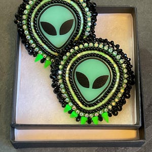 K&ouml;nnte beinhalten: Ein Paar handgefertigte Ohrringe mit Alien-Motiv. Jeder Ohrring zeigt ein hellgr&uuml;nes Alien-Gesicht mit schwarzen Augen, umgeben von schwarzen und irisierenden Perlen. Die Ohrringe haben einen neongr&uuml;nen Fransenbesatz und werden in einer schwarzen Box pr&auml;sentiert.