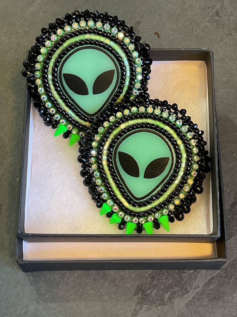 K&ouml;nnte beinhalten: Ein Paar handgefertigte Ohrringe mit Alien-Motiv. Jeder Ohrring zeigt ein hellgr&uuml;nes Alien-Gesicht mit schwarzen Augen, umgeben von schwarzen und irisierenden Perlen. Die Ohrringe haben einen neongr&uuml;nen Fransenbesatz und werden in einer schwarzen Box pr&auml;sentiert.