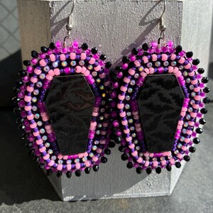 May include: A pair of dangle earrings featuring a central black coffin-shaped charm with a subtle bat pattern. The charms are surrounded by concentric rows of pink, purple, and iridescent beads, finished with a border of small black beads.