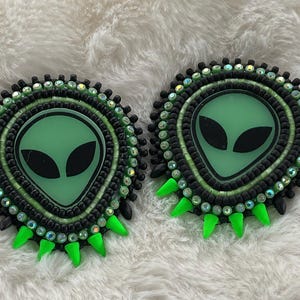 May include: Pair of handmade alien-themed earrings. Each earring features a green alien face with black eyes, surrounded by black and green beads. The design includes green spikes at the bottom and iridescent accents, set against a white, fluffy background.