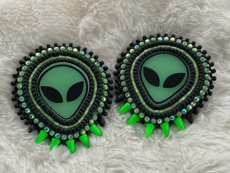 May include: Pair of handmade alien-themed earrings. Each earring features a green alien face with black eyes, surrounded by black and green beads. The design includes green spikes at the bottom and iridescent accents, set against a white, fluffy background.