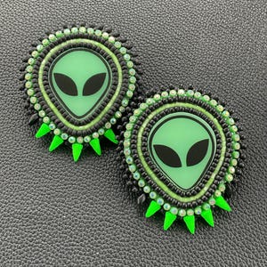 May include: Two round, beaded alien-themed earrings. Each earring features a green alien face with black eyes, surrounded by black and green beads. The earrings have green, triangular accents around the edges.
